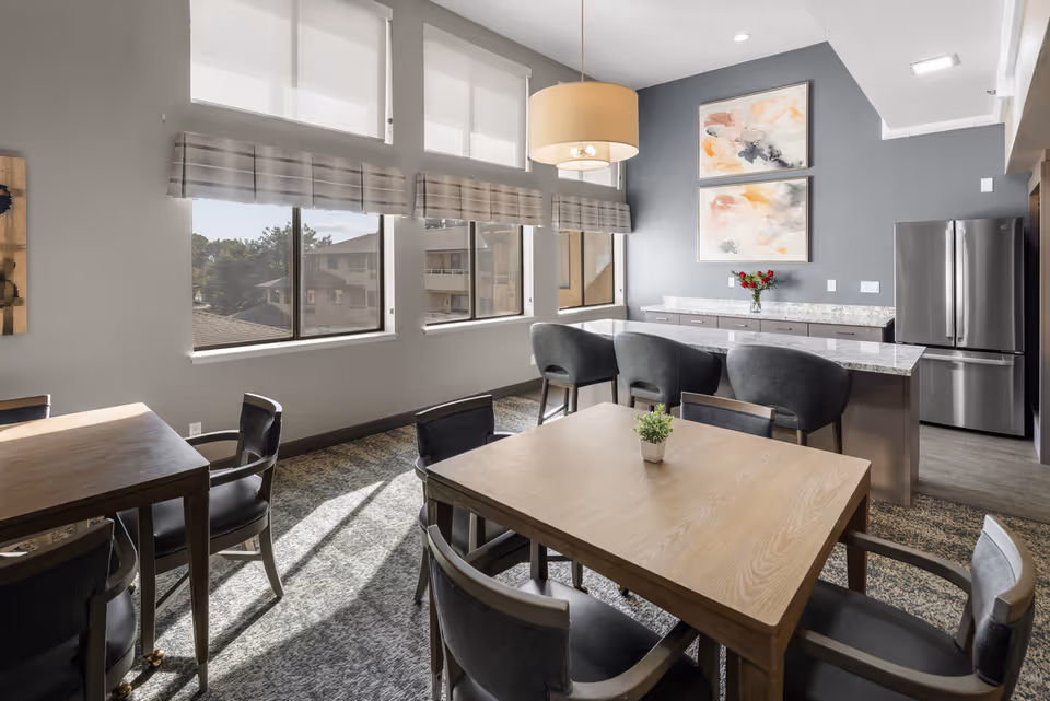 A bright dining area in a senior living community featuring wooden tables with black cushioned chairs, a kitchen island with three gray upholstered bar stools, a stainless steel refrigerator, and two abstract paintings on a gray accent wall. Large windows with white roller shades and plaid valances allow natural light to fill the room.