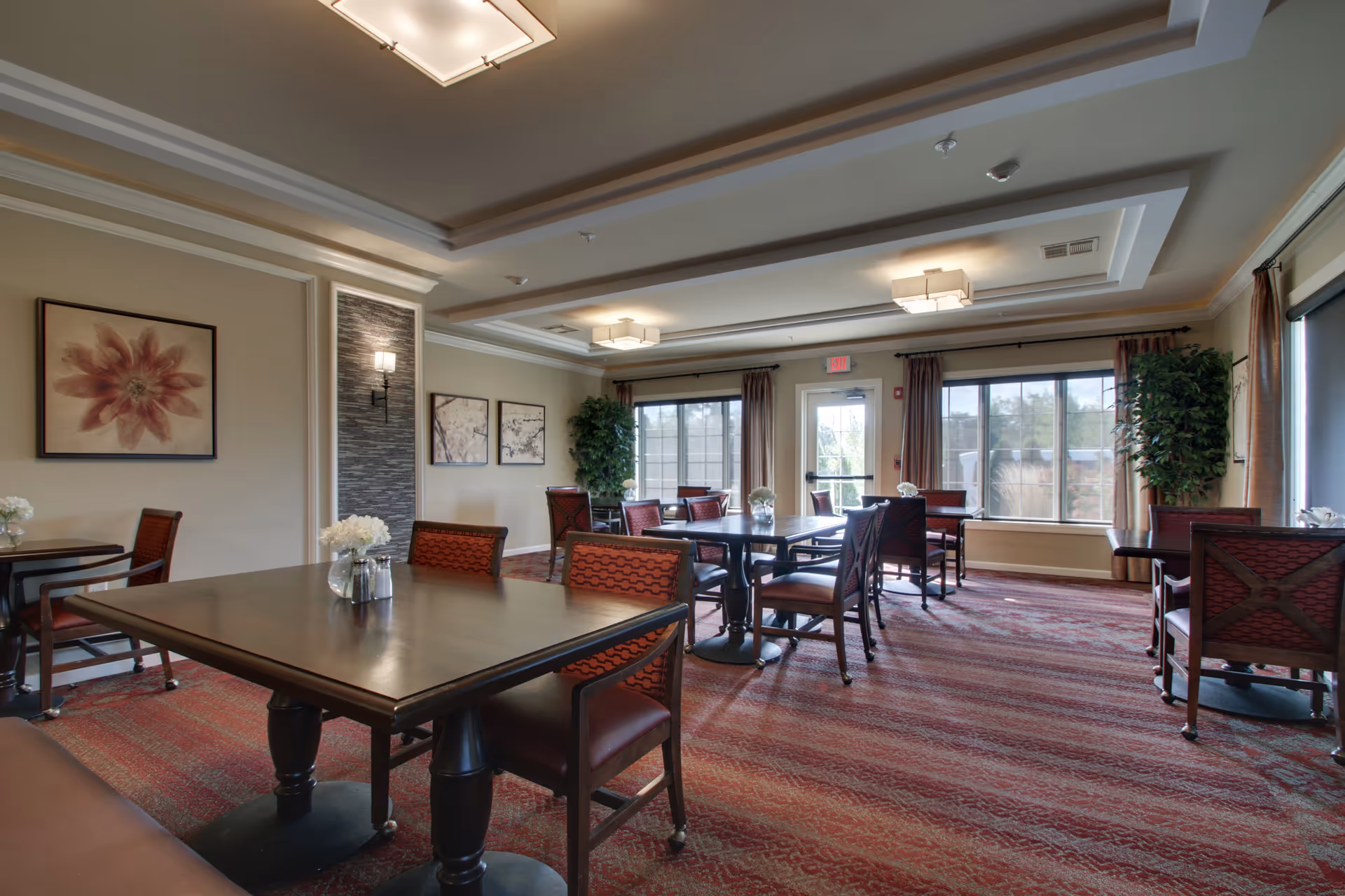 A well-lit dining room in Evergreen Senior Living with multiple dark wooden tables and red cushioned chairs. The room features large windows with curtains, framed artwork on the walls, a patterned carpet, and ceiling lights. There are also decorative plants near the windows.