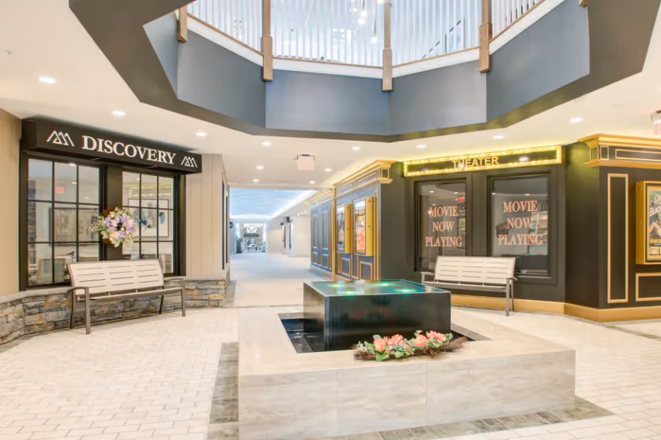 Interior view of a senior living facility hallway featuring a central water fountain with flowers around it, benches on either side, and entrances labeled 'DISCOVERY' and 'THEATER' with signs indicating 'MOVIE NOW PLAYING'. The area is well-lit with a modern design and a second-floor balcony above.