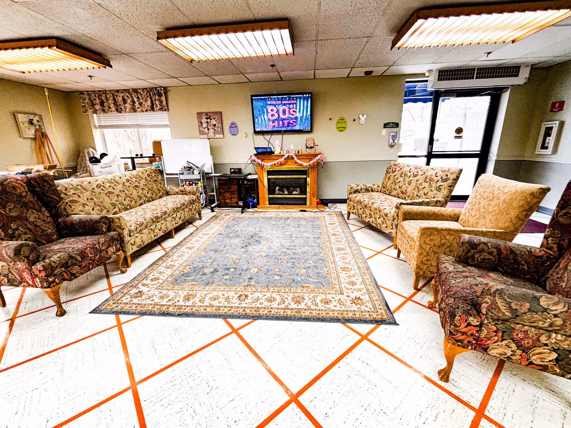 A cozy living room area with patterned armchairs and sofas arranged around a large decorative rug. A TV mounted on the wall displays 'World's Greatest 80s Hits'. Below the TV is a fireplace with a garland decoration. The room has a tiled floor with orange grid lines, fluorescent ceiling lights, and a glass door leading outside.