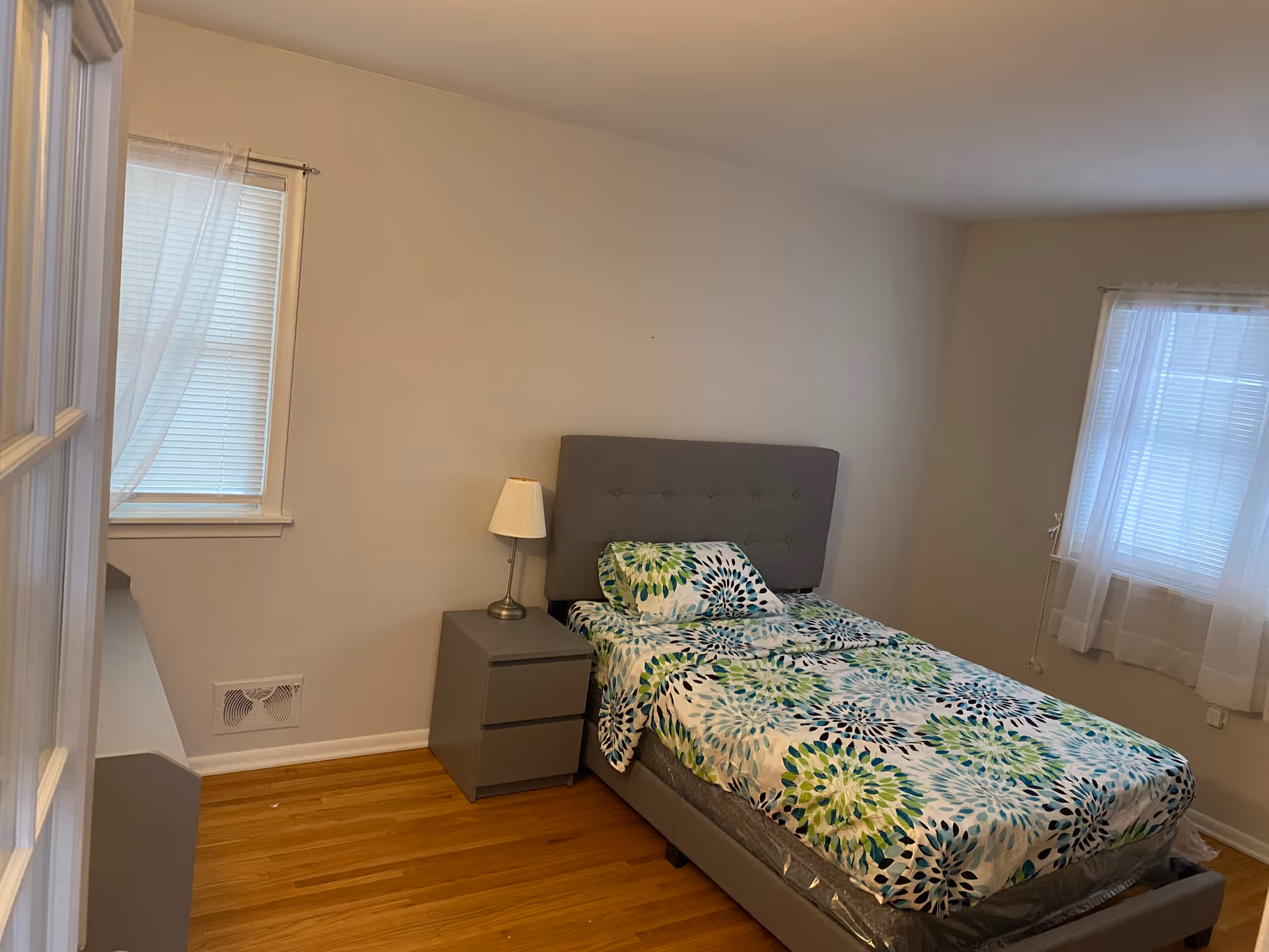 A simple bedroom with a single bed featuring a gray upholstered headboard and a colorful floral bedspread in shades of green, blue, and white. Next to the bed is a gray nightstand with a small lamp. The room has wooden flooring and two windows with white blinds and sheer curtains, allowing natural light to enter.