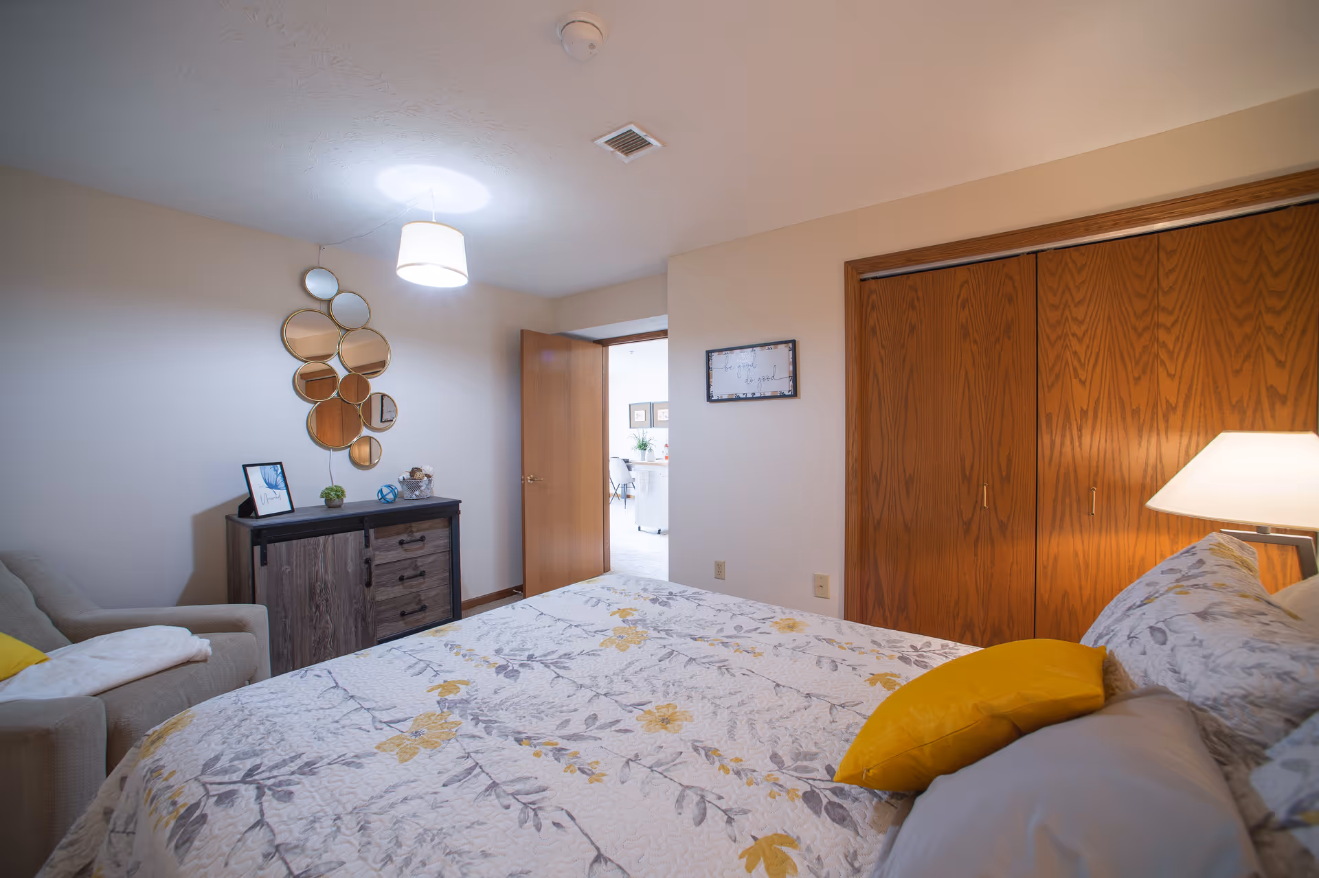 A bedroom with a bed covered in a floral patterned quilt with yellow and gray flowers. There is a yellow pillow and a gray pillow on the bed. To the right of the bed is a wooden closet with double doors. A lamp with a white shade is on a nightstand next to the bed. On the left side of the room, there is a gray armchair with a white blanket draped over one arm. Next to the armchair is a dark wooden dresser with a decorative mirror made of multiple circular mirrors above it. The door to the room is open, showing a glimpse of another room with a white floor and some furniture.