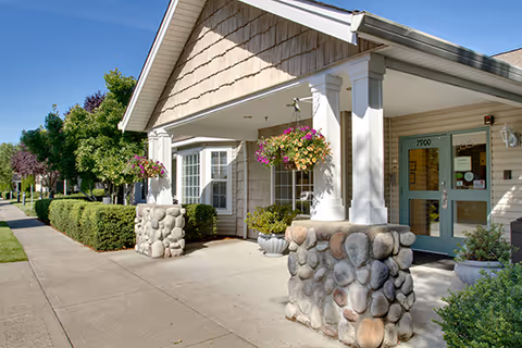 Exterior view of the entrance to a senior living facility with a covered porch supported by stone pillars, hanging flower baskets, and a pathway lined with bushes and trees.