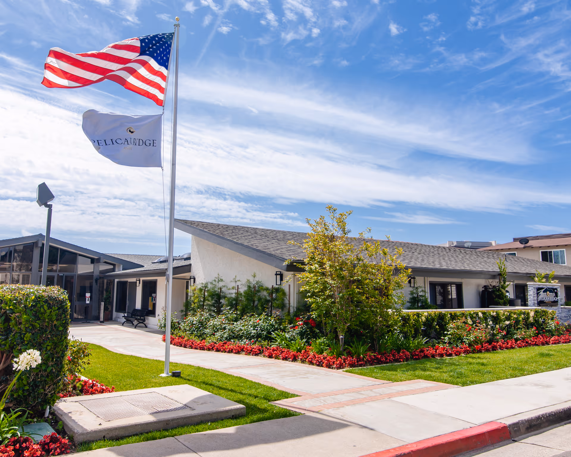 Exterior view of Pelican Ridge Post-Acute facility with a well-maintained garden featuring red flowers and green shrubs. Two flagpoles display the American flag and a white flag with the Pelican Ridge logo. The building has a light-colored facade and a sloped roof under a partly cloudy blue sky.