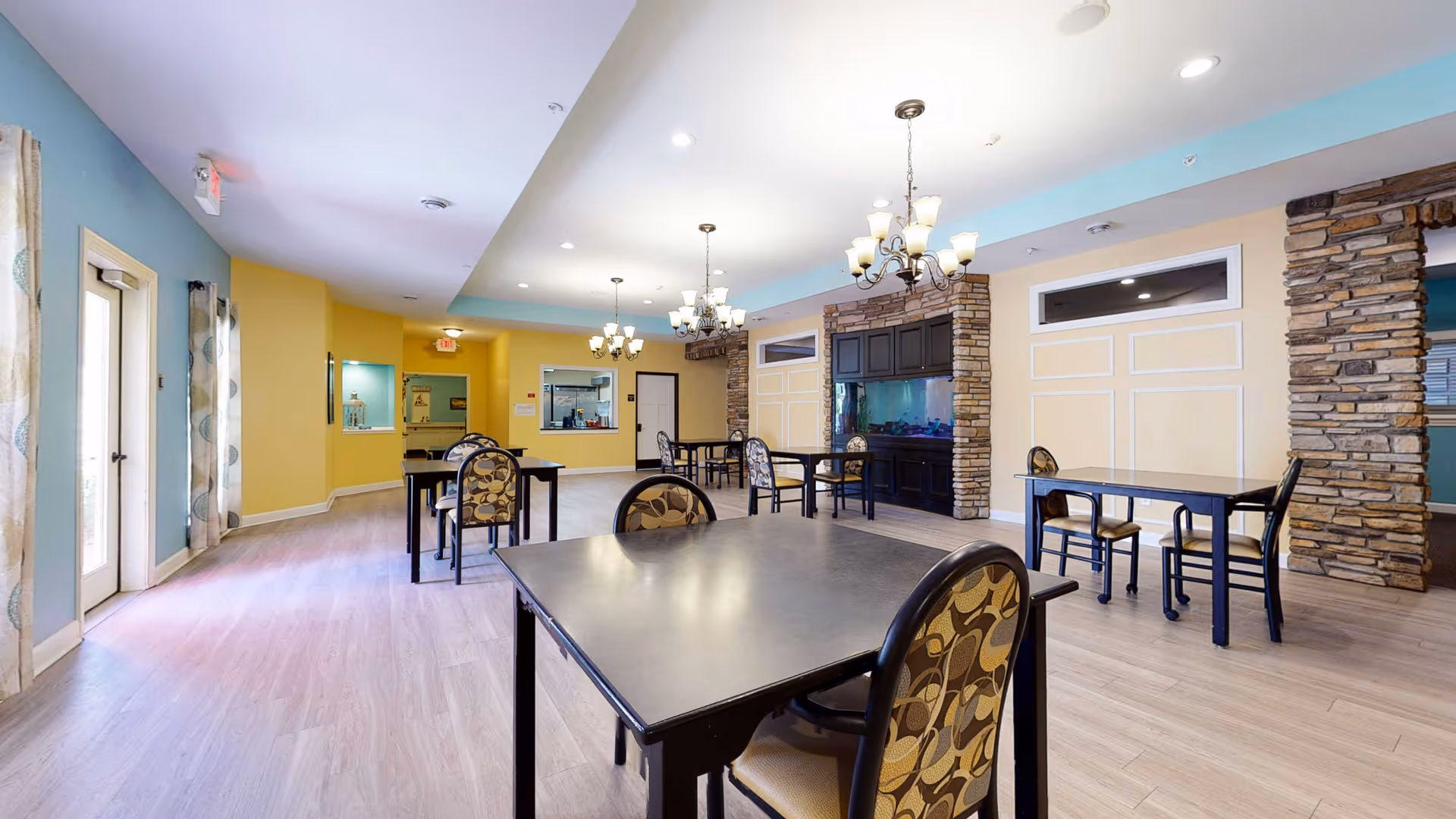 A spacious dining room in a retirement living facility with several black tables and patterned chairs. The room features light wood flooring, yellow and blue walls, stone pillars, and multiple chandeliers hanging from the ceiling. There is a large built-in aquarium on one wall and windows with curtains on the left side.