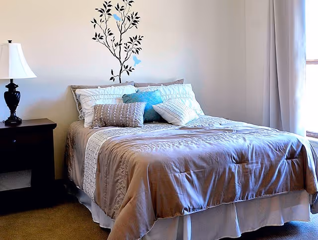 A neatly made bed with beige and white bedding and multiple pillows in a bedroom. There is a black bedside table with a black lamp on it to the left of the bed. On the wall above the bed is a decorative decal of a tree with blue birds. A window with light curtains is on the right side, allowing natural light into the room.