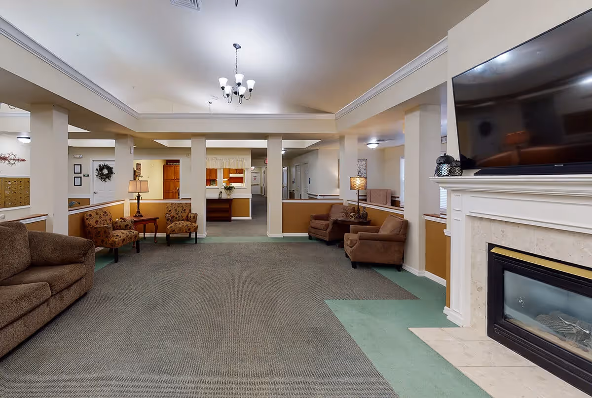 A spacious common area in a senior living facility with multiple seating arrangements including brown sofas and armchairs. There is a fireplace with a large flat-screen TV mounted above it on the right side. The room has beige walls with white trim, carpeted floors, and several lamps providing warm lighting. In the background, there is a reception desk and a hallway leading to other rooms.
