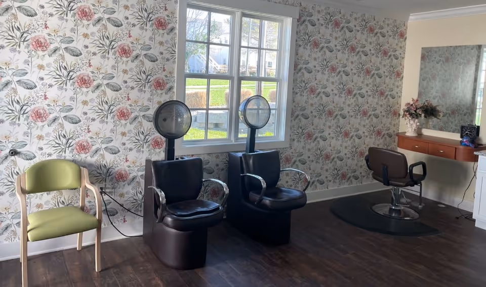 Interior view of a hair salon area with two black hair drying chairs and one styling chair in front of a mirror. The walls are decorated with floral wallpaper, and there is a window letting in natural light. A green waiting chair is also visible.
