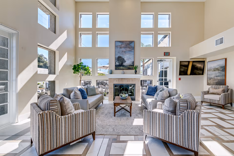 A bright and spacious senior living facility common area with high ceilings and multiple square windows letting in natural light. The room features a central fireplace with a painting above it, flanked by two sofas with blue and gray cushions, two striped armchairs, a wooden coffee table on a light-colored rug, and a flat-screen TV mounted on the wall. There is also a potted plant near the windows and an exit door leading outside to a patio area with tables and umbrellas.