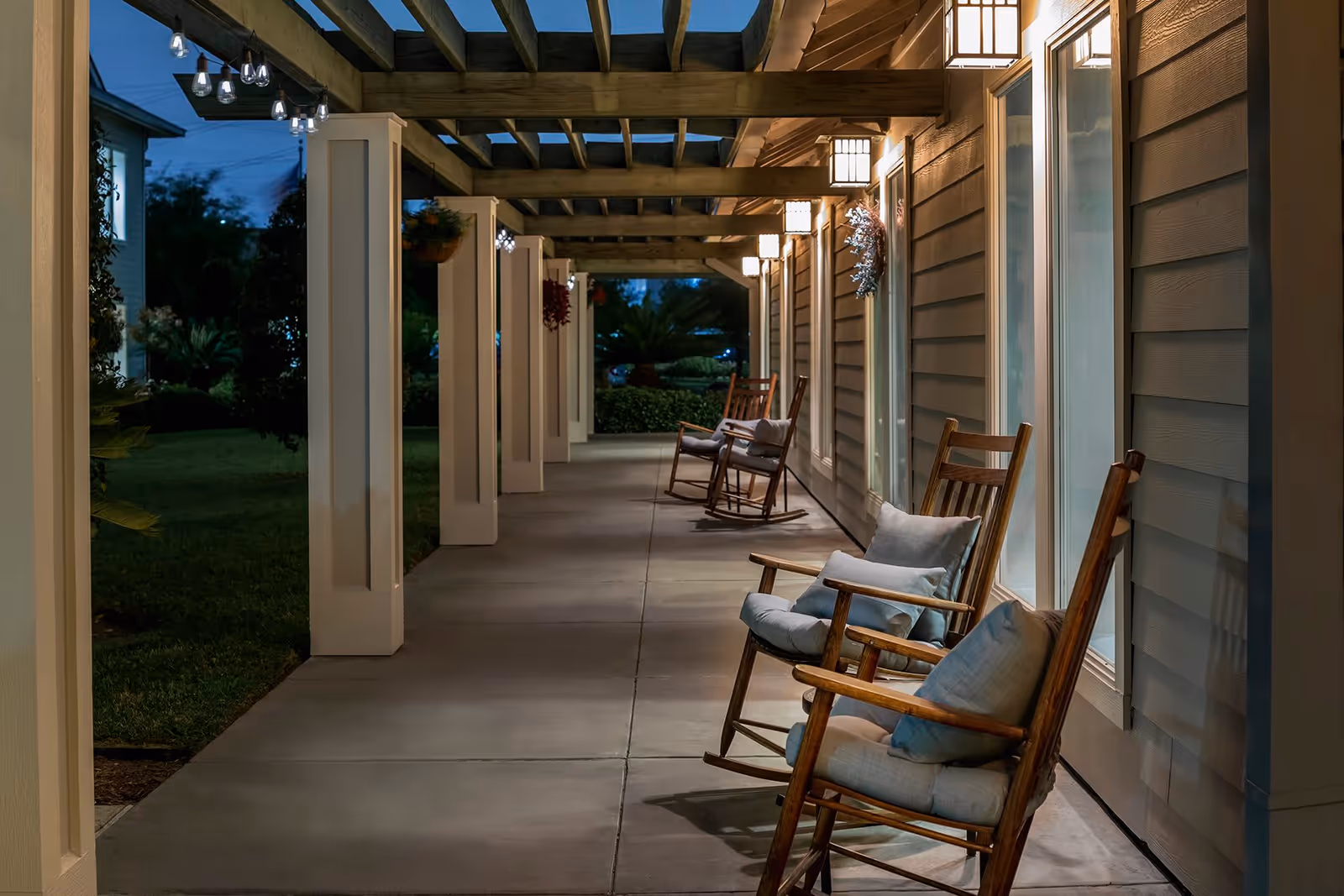 A covered outdoor porch area at night with wooden rocking chairs lined up along the wall of a building. The porch has a wooden pergola overhead with hanging lights, and the area is softly illuminated by wall-mounted lantern-style lights. There is greenery visible beyond the porch.