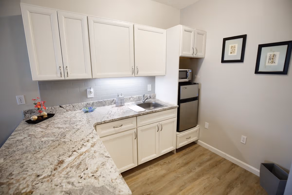 A small kitchen area with white cabinets, a granite countertop, a stainless steel sink, a mini refrigerator, and a microwave. The walls are light-colored with two framed pictures hanging on one side. The floor is wood-style laminate.