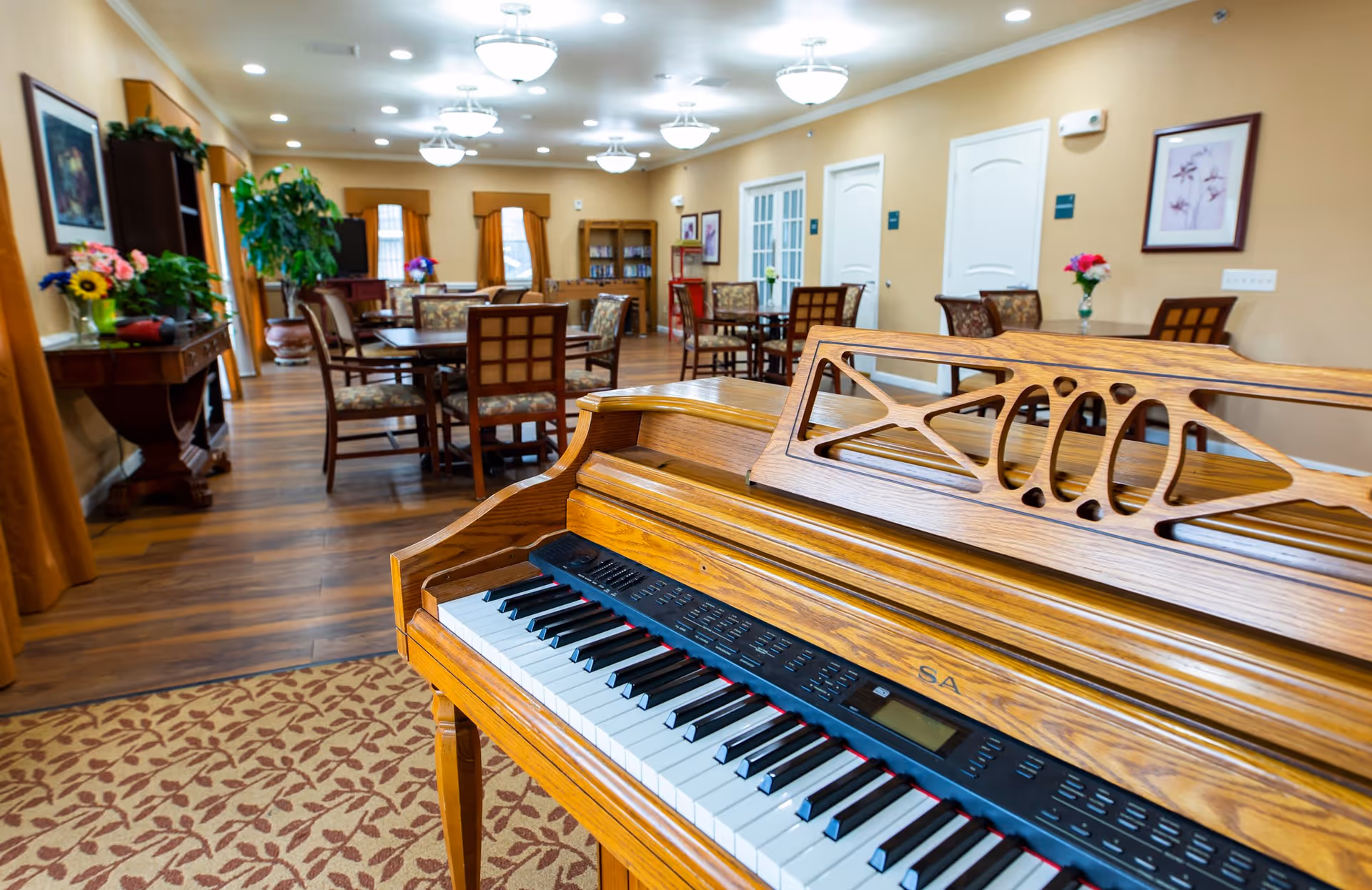Interior view of a senior living facility common area featuring a wooden piano in the foreground, several tables and chairs arranged throughout the room, decorative plants, framed artwork on the walls, and warm lighting from ceiling fixtures.