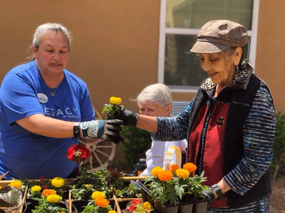 Two women gardening outdoors, one wearing a blue shirt with 'Legacy at Santa Fe' on it handing a yellow flower to the other woman wearing a hat and a patterned vest. A third person is seated in the background near a window.
