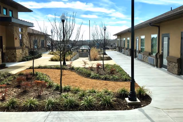 Courtyard walkway between single-story beige buildings with landscaped planting beds, young trees and lamp posts under a blue sky.