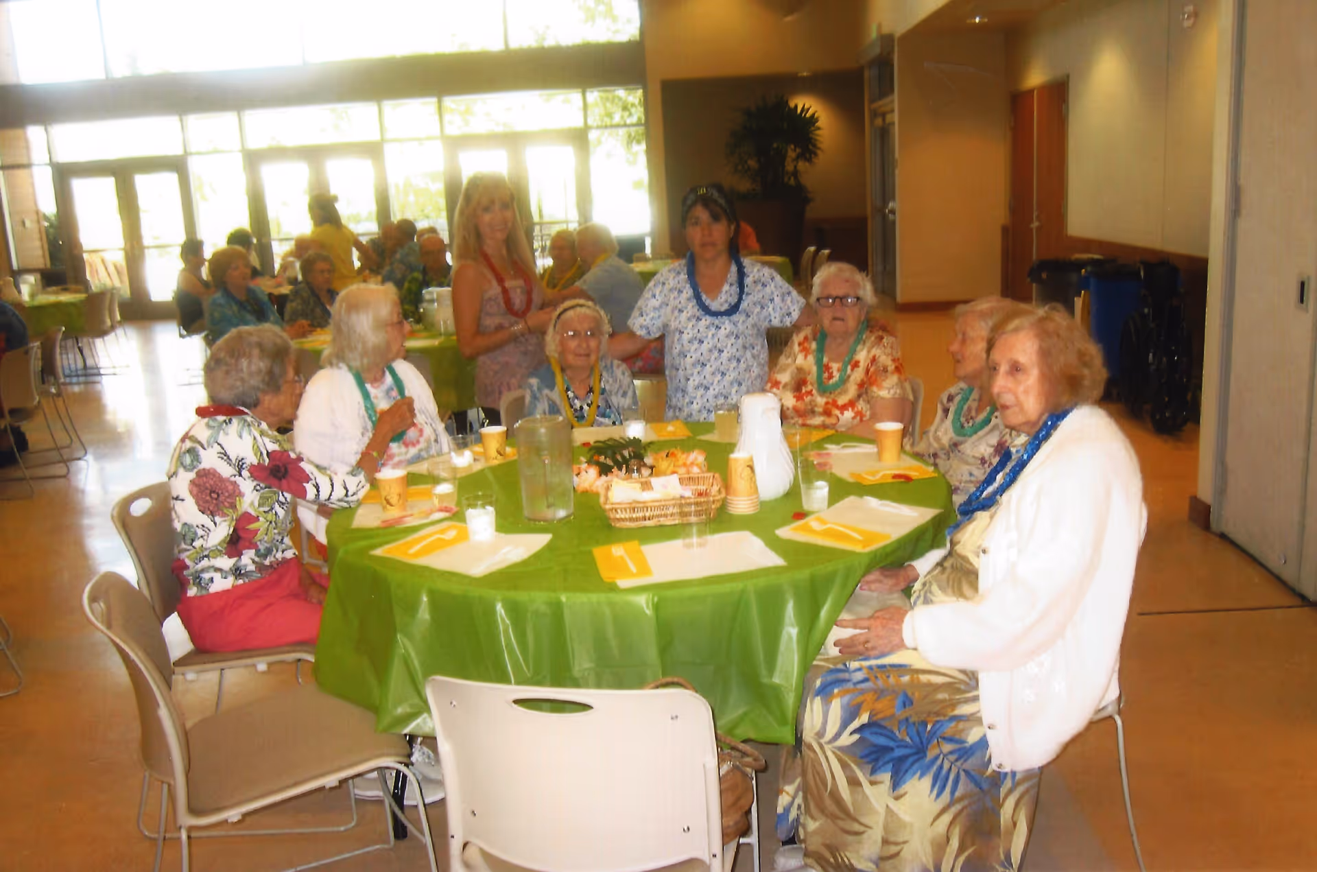 A group of elderly women sitting around a round table covered with a green tablecloth in a well-lit dining area. The table has cups, napkins, and a basket with food items. Two women are standing behind the seated group, and other people are visible in the background near large windows.