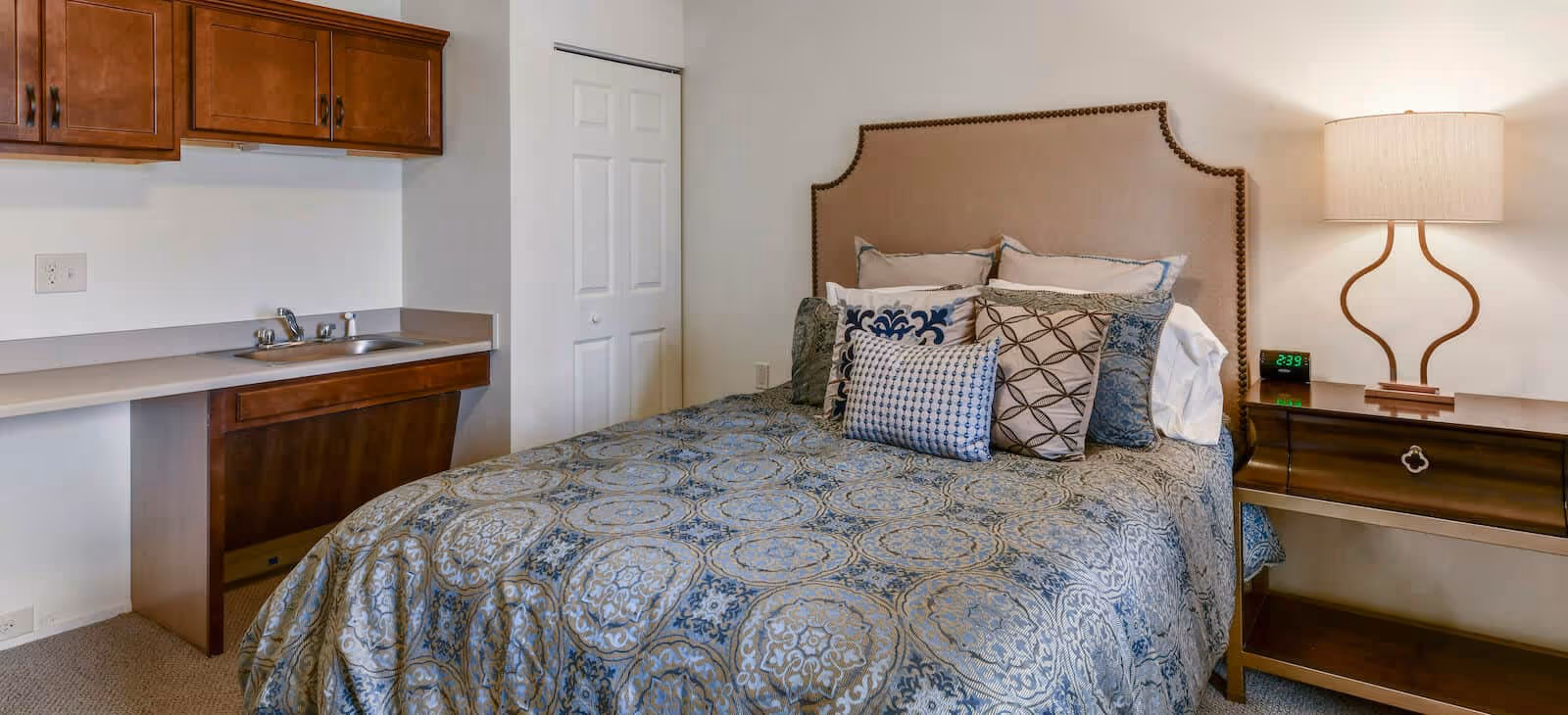 A furnished bedroom with a patterned bed and upholstered headboard, a nightstand and lamp, and a small sink with cabinets along one wall.