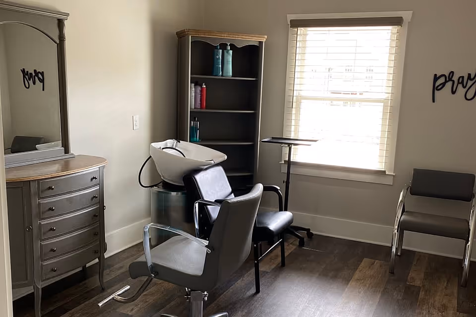 A small salon area inside a senior living facility with a hair washing station, two salon chairs, a wooden dresser with a mirror, a shelving unit with hair care products, and a window with blinds letting in natural light. The word 'pray' is visible on the wall.