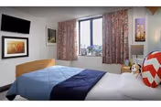 A bedroom with a bed covered in a blue and navy quilt, a white pillow with a red chevron pattern, and a small stuffed dog toy. The room has a window with floral curtains, a wall-mounted flat screen TV, and framed artwork on the walls.