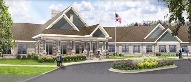 Exterior view of a single-story senior living facility building with a covered entrance, stone and siding facade, landscaped greenery, an American flag on a flagpole, and people walking and using a mobility scooter near the entrance.