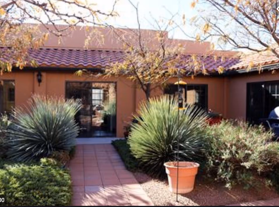 Outdoor view of a courtyard at Carefree Assisted Living Center featuring a tiled walkway leading to a building entrance with large glass doors. The courtyard is landscaped with various desert plants, including spiky green shrubs and a potted plant, under leafless tree branches.
