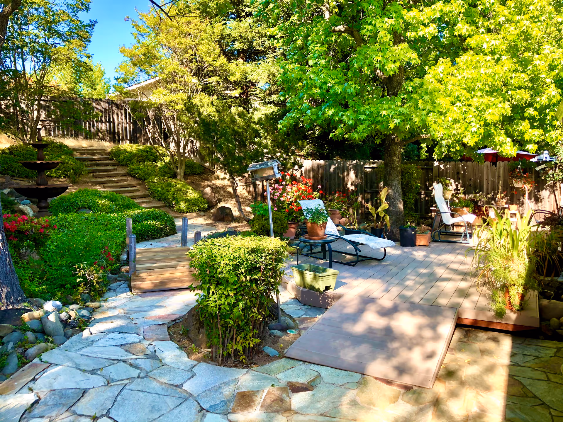 A sunny outdoor garden area with a stone pathway leading to wooden steps and a small wooden bridge. There are green bushes, trees, and various potted plants around a wooden deck with two reclining chairs. A wooden fence encloses the garden.