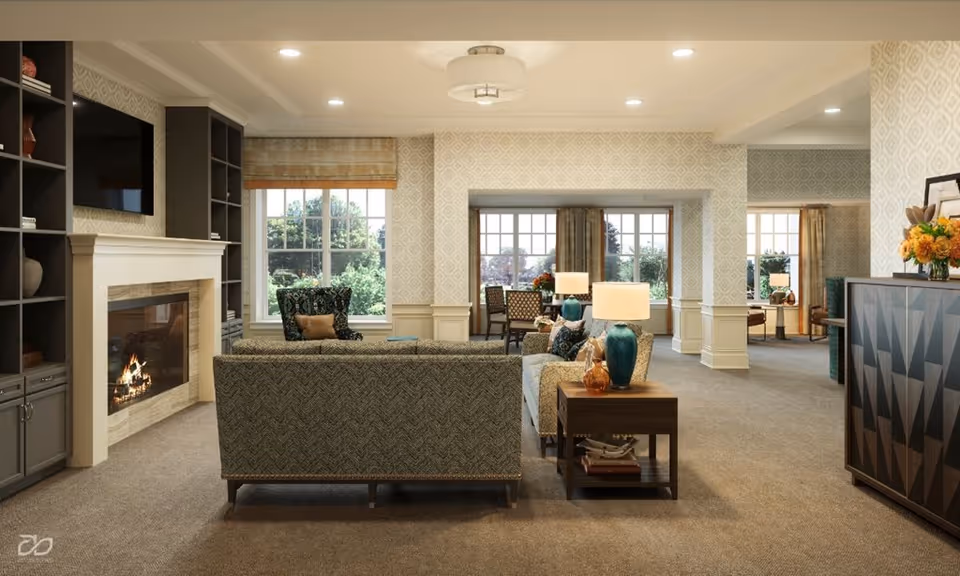 A cozy living room area with a lit fireplace, a mounted TV above it, and built-in shelves on the left. There are two sofas facing the fireplace, a side table with two blue lamps and decorative items. Large windows with beige curtains allow natural light to fill the room. The walls have a patterned wallpaper and the carpet is beige. In the background, there is a dining area with chairs and a table near more windows.
