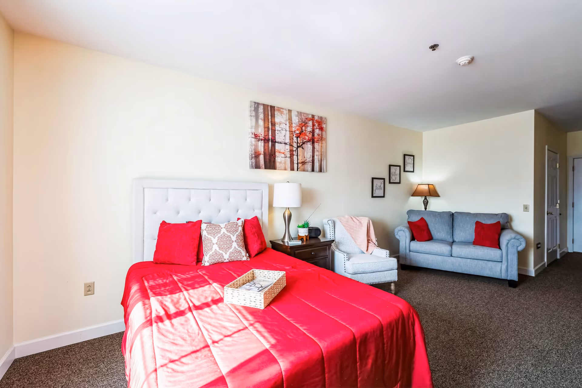 A senior living facility bedroom featuring a bed with a red bedspread and decorative pillows, a nightstand with a lamp and small plant, a light gray armchair with a pink throw blanket, and a light blue sofa with red pillows. The walls are decorated with framed artwork and a painting of trees with red leaves.