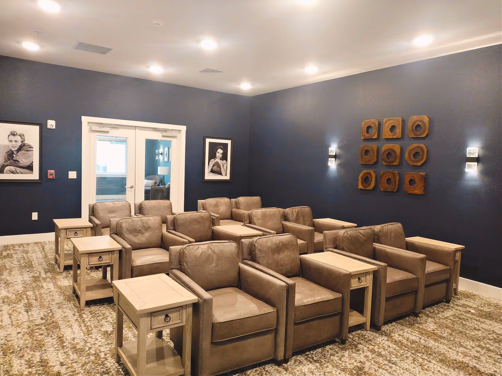 A cozy seating area with three rows of brown leather armchairs arranged in pairs, each pair separated by a light wooden side table. The room has dark blue walls decorated with black and white framed portraits and a wooden wall art piece consisting of nine square blocks with circular cutouts. The floor is carpeted with a patterned design, and there are double glass doors at the back of the room.