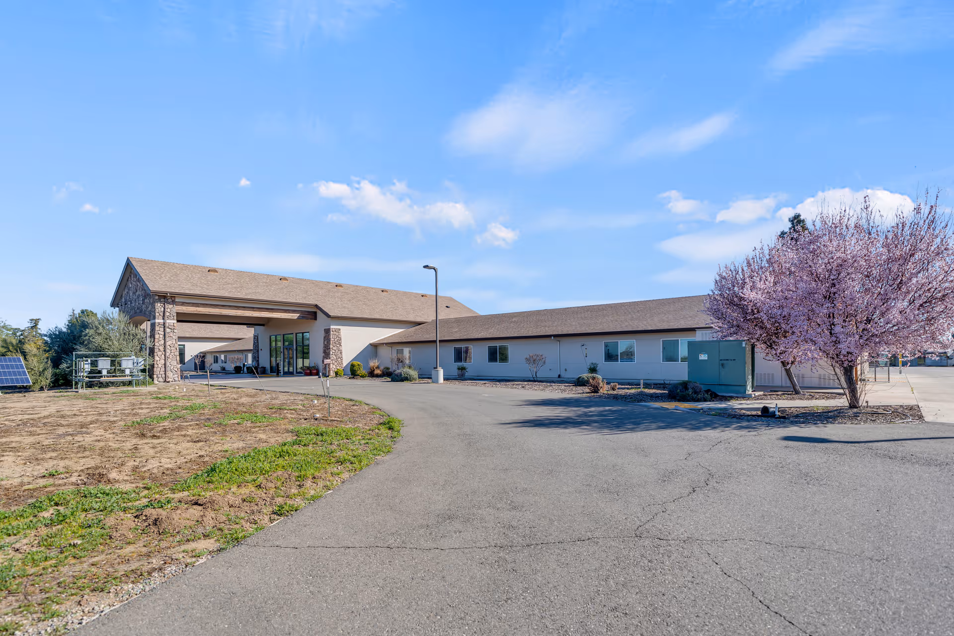 Exterior view of a single-story building with a covered entrance, stone pillars, and a paved driveway. There is a blooming tree with pink flowers on the right side and some greenery on the left side under a blue sky with scattered clouds.