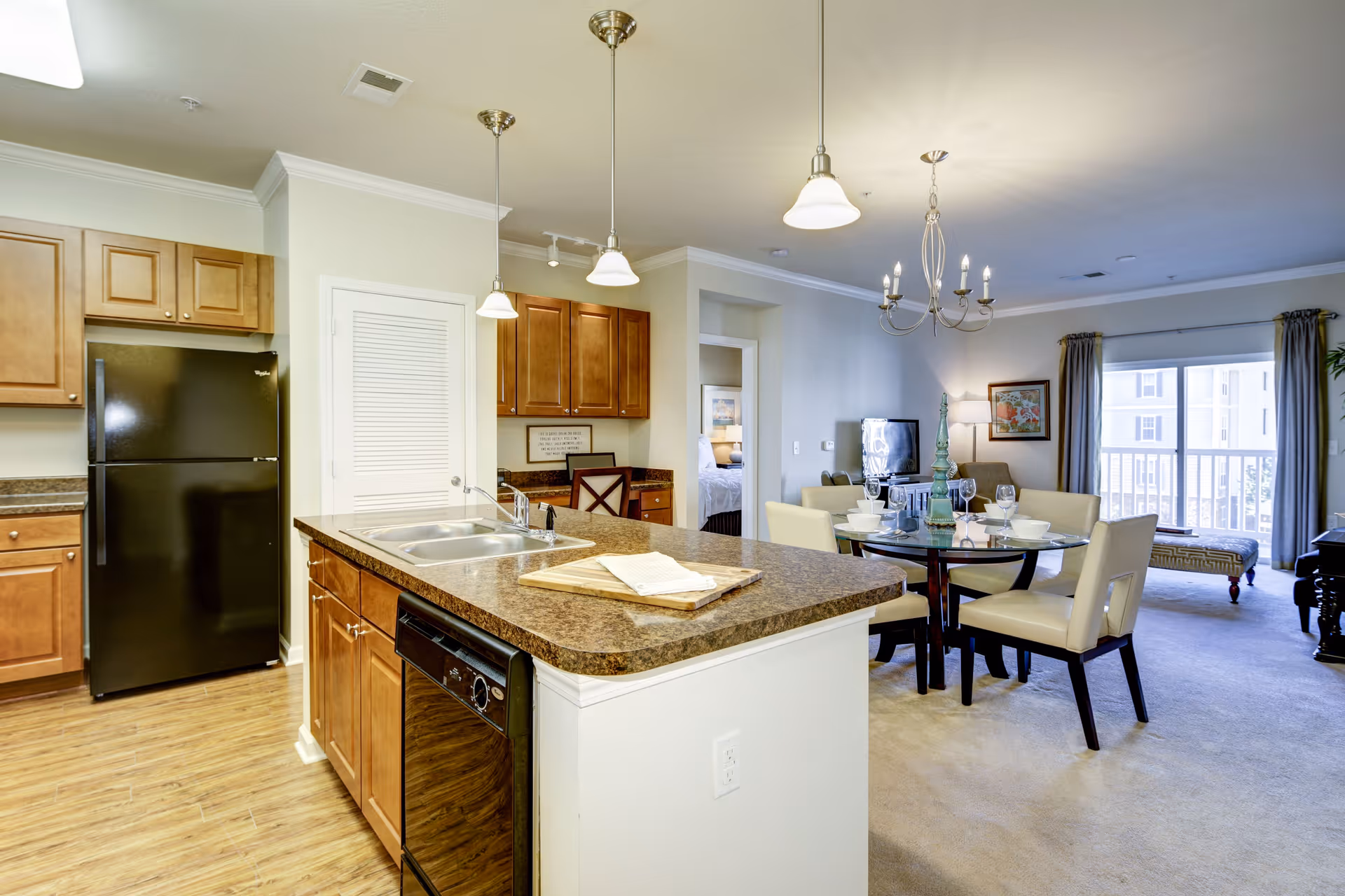 Open kitchen and dining area in a senior living facility. The kitchen features wooden cabinets, a black refrigerator, a double sink on a brown countertop island with a dishwasher underneath, and pendant lights hanging above. The dining area has a round glass table set with plates, glasses, and a decorative centerpiece, surrounded by cream-colored chairs. In the background, there is a living room with a TV, a framed picture on the wall, a floor lamp, and a sliding glass door leading to a balcony with curtains.