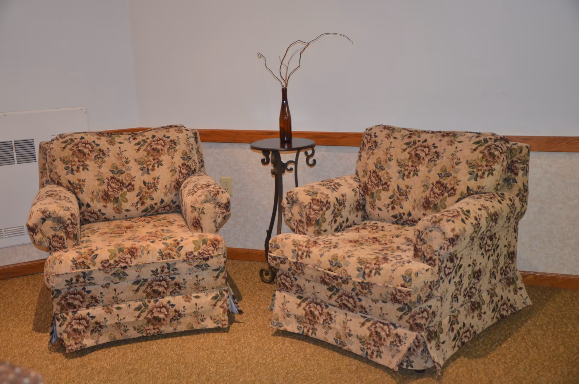 Two floral upholstered armchairs with a small round side table between them holding a brown vase with dried branches, set against a beige wall with wooden trim and carpeted floor.