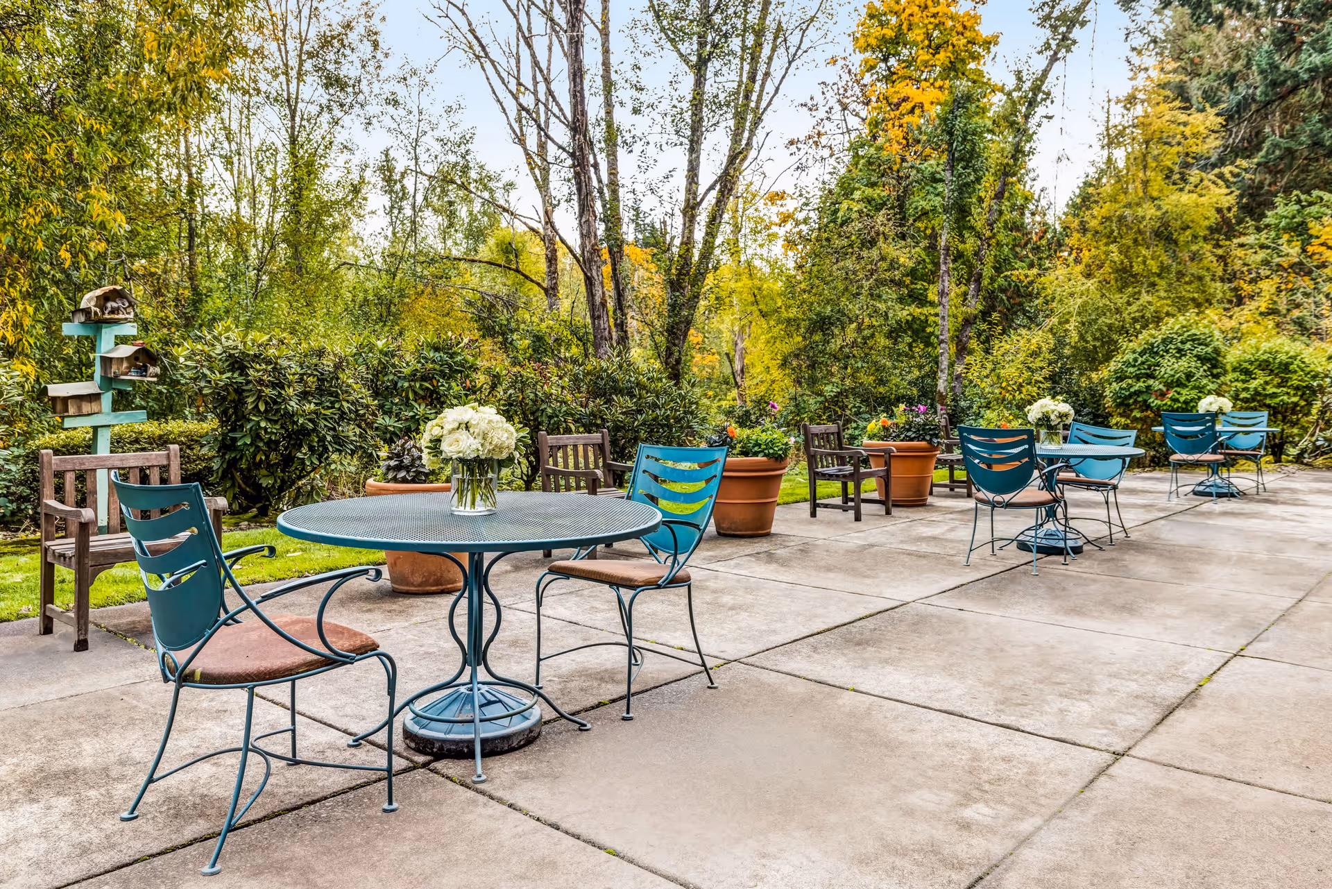 Outdoor patio area with several round metal tables and chairs arranged on a concrete surface. Each table has a vase with white flowers. Large potted plants and greenery surround the patio, with tall trees and bushes in the background.