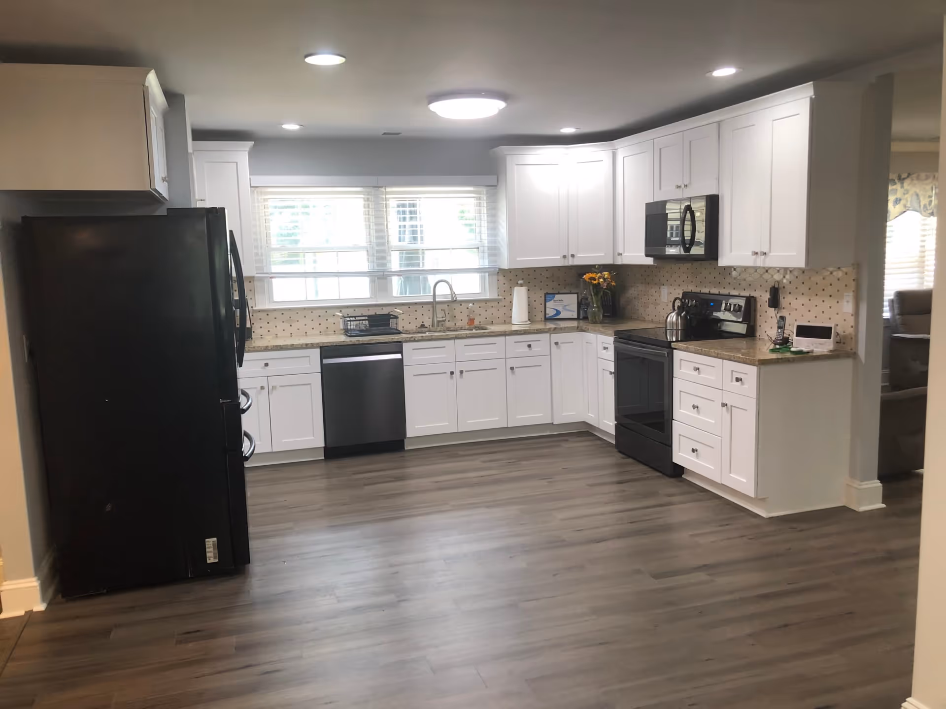 A modern kitchen with white cabinets, a black refrigerator, black dishwasher, black stove, and black microwave. The kitchen has a light-colored countertop and a tiled backsplash. There is a window above the sink letting in natural light, and the floor is a wood-like laminate.