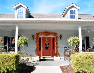 Front exterior view of a light gray building with a red door decorated with autumn-themed garlands and pumpkins. The porch has two hanging plants, two black rocking chairs, and two ceiling fans. There are bushes and a walkway leading to the entrance under a clear blue sky.