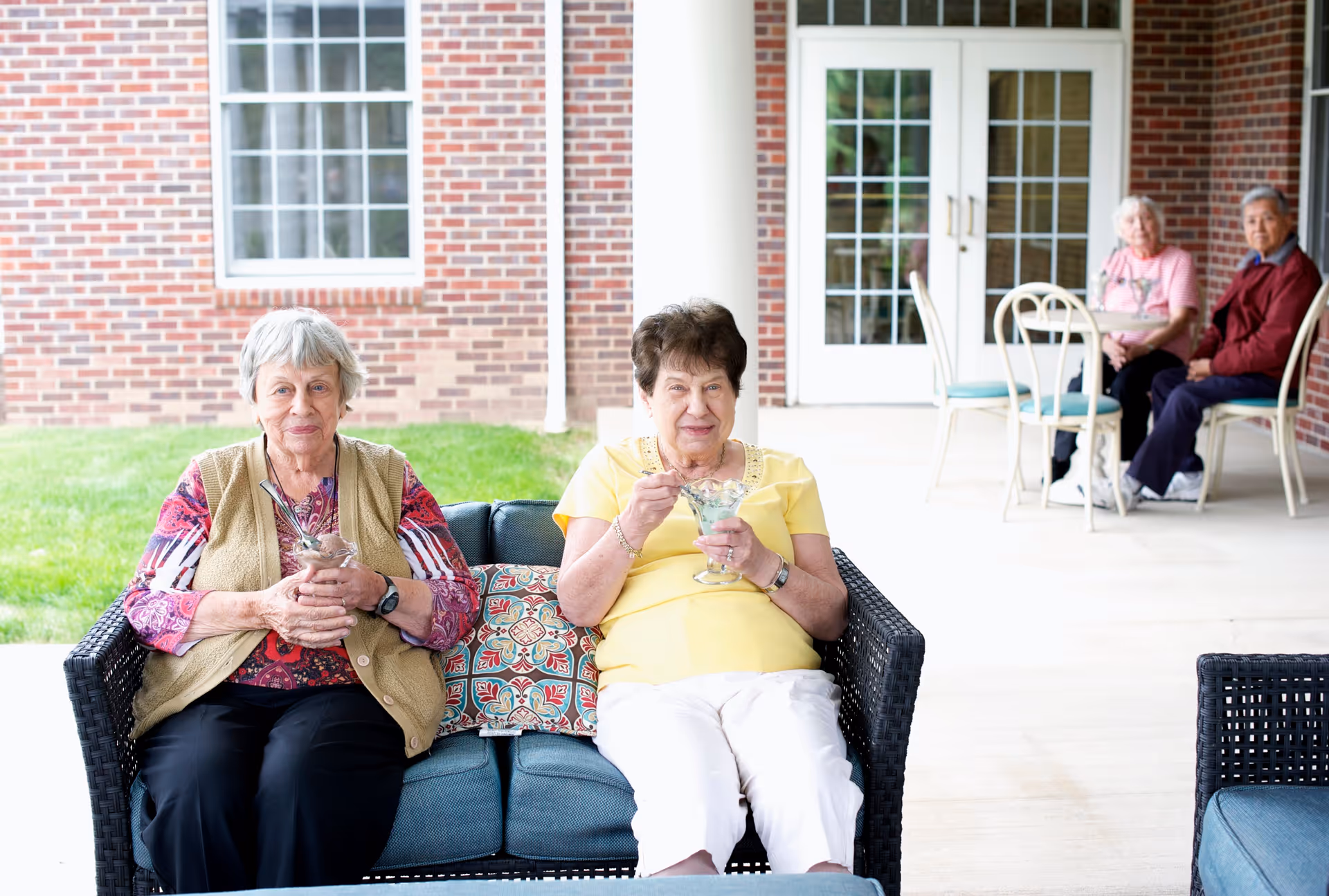 Two elderly women sitting on a cushioned outdoor wicker sofa, enjoying ice cream or dessert. Behind them, two other elderly individuals sit at a small round table with chairs on a covered patio area outside a brick building with large windows and glass doors.