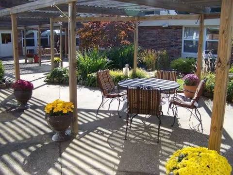 Outdoor patio with round table, six chairs, and potted flowers under a wooden pergola.