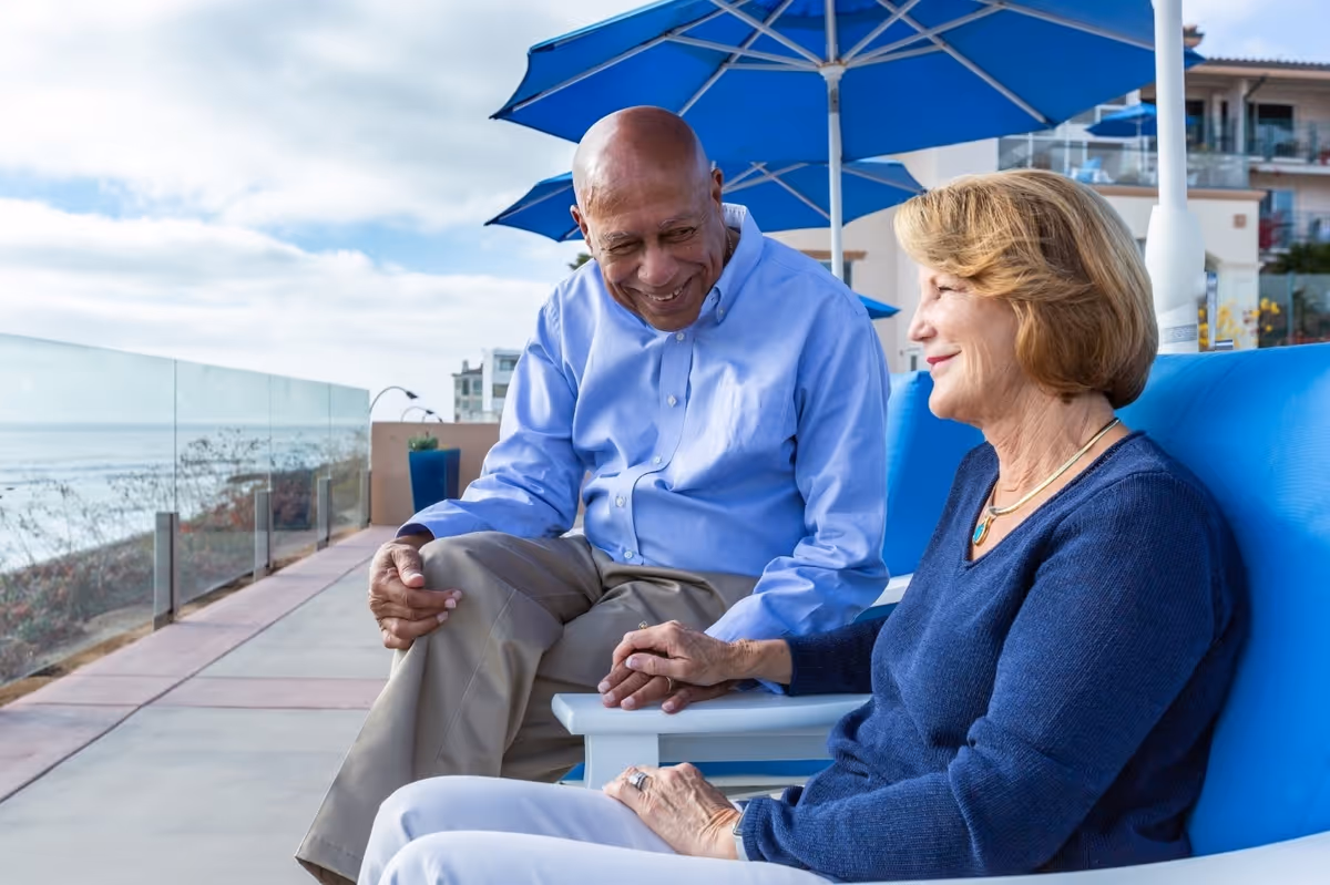 An elderly man and woman sitting outdoors on blue chairs under blue umbrellas, holding hands and smiling at each other. They are on a paved patio area with a glass railing overlooking the ocean and some buildings in the background.