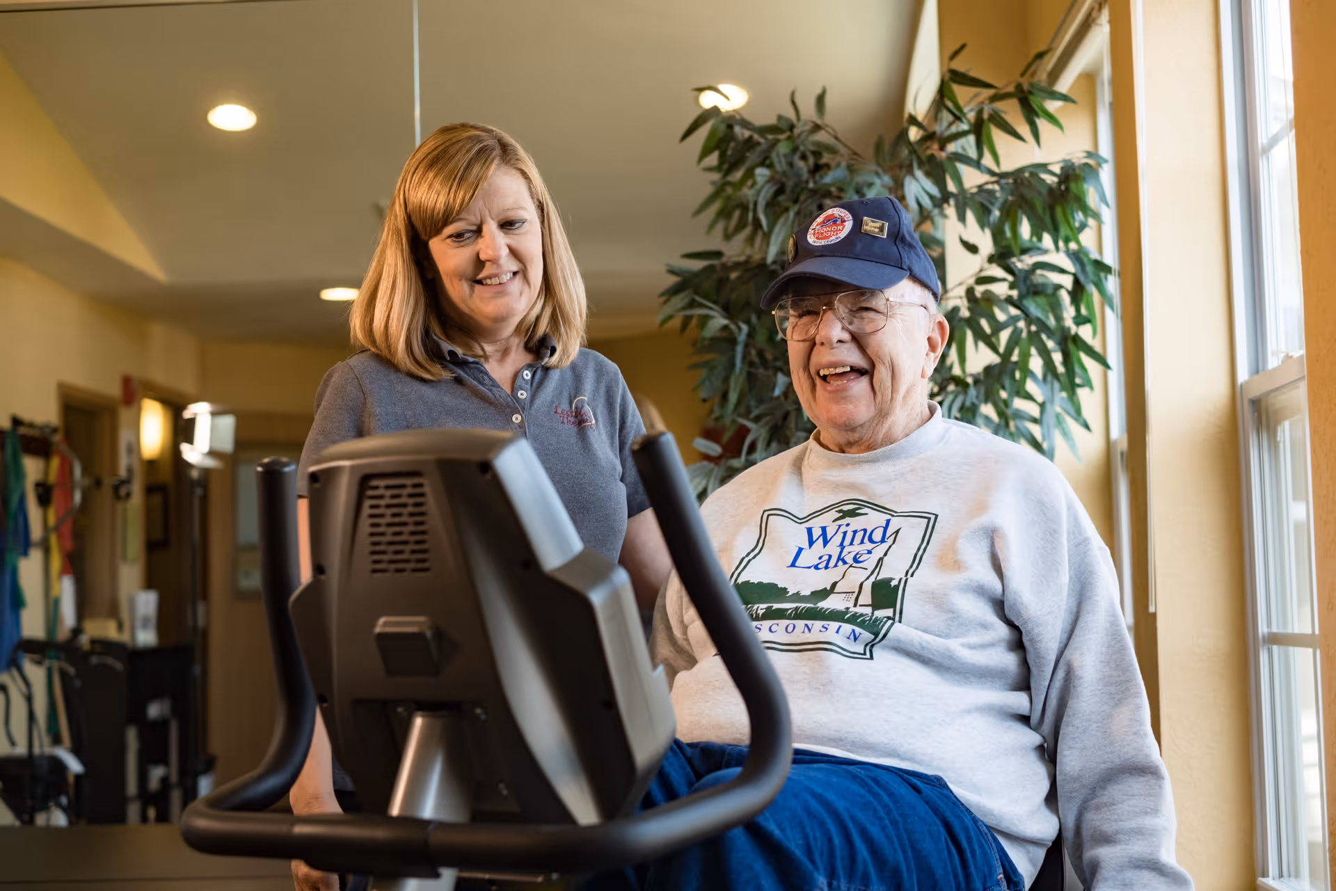 An elderly man wearing a cap and a sweatshirt with 'Wind Lake Wisconsin' printed on it is smiling while using a stationary exercise bike indoors. A woman, possibly a caregiver or fitness instructor, stands beside him smiling and offering support. The room has warm lighting and a large plant near a window.
