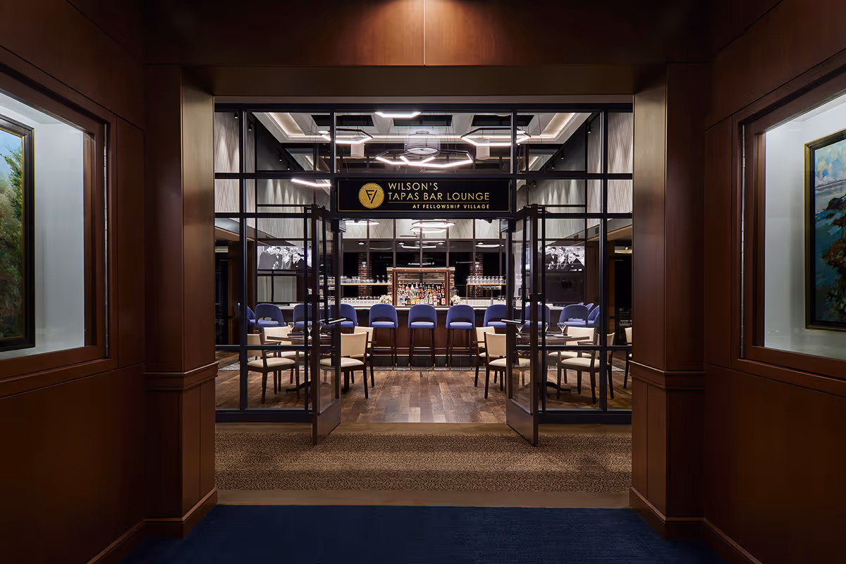 Entrance view into Wilson's Tapas Bar Lounge showing a bar counter with stools and dining tables framed by a wood-paneled corridor.