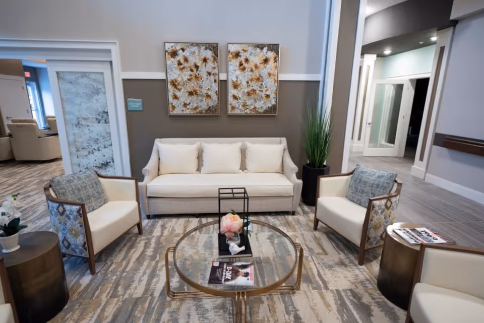 A modern living room area with a white sofa and two armchairs with patterned upholstery. There is a round glass coffee table with a flower arrangement and magazines on it. The room has a neutral color palette with beige and gray tones, two floral paintings on the wall, and a tall green plant in a black pot. The flooring is a patterned carpet, and there are doorways leading to other rooms in the background.