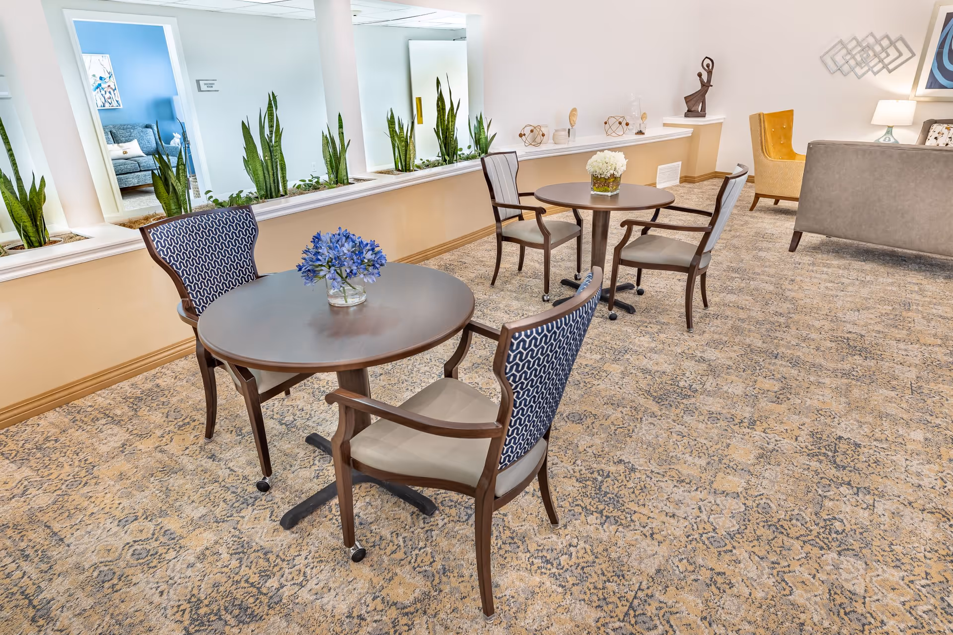 A bright and inviting common area in a senior living facility with two round tables, each surrounded by two chairs. One table has a small vase with blue flowers. The room features a patterned carpet, a row of tall green plants along a half wall with mirrors behind them, and a seating area with a gray sofa and a yellow armchair. Decorative items and artwork adorn the walls and surfaces.