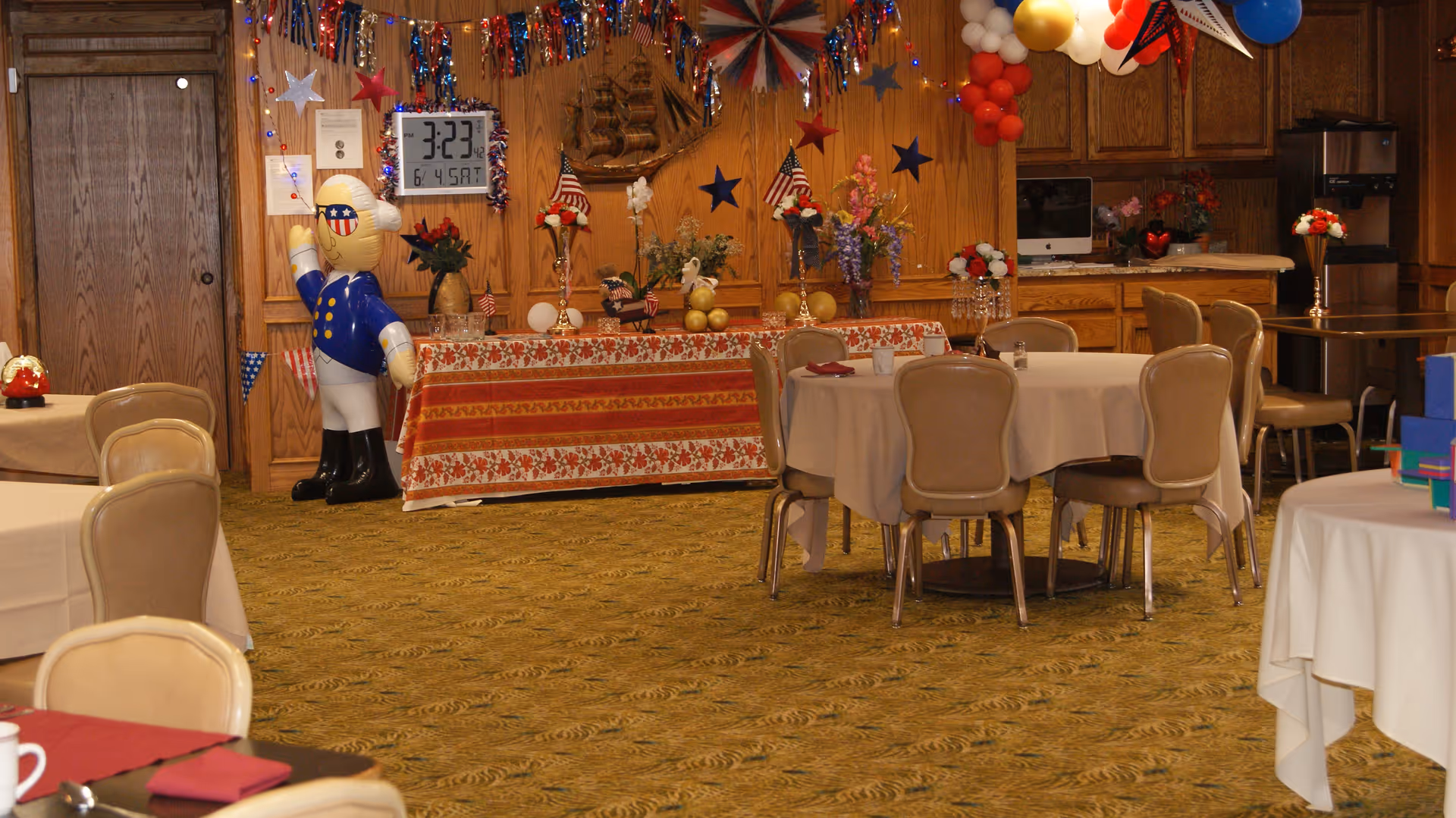 Dining room with round tables and chairs, an American-themed decorated buffet table, balloons, and a large inflatable sailor figure.
