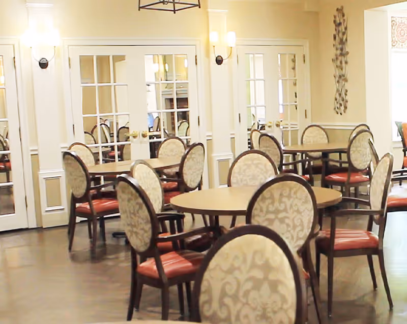 A dining room with multiple round tables and chairs featuring patterned upholstery and wooden frames. The room has light-colored walls, wall sconces, and glass-paneled double doors in the background.