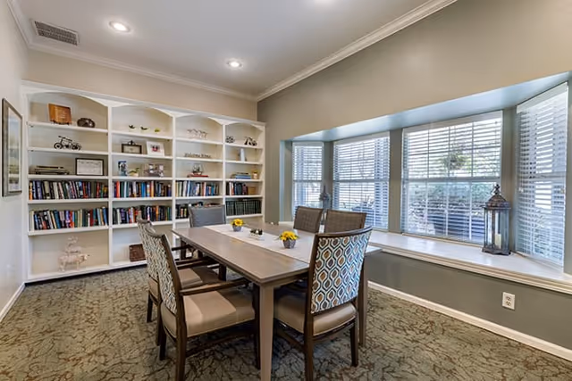 A well-lit room with a rectangular wooden table surrounded by six upholstered chairs. Behind the table is a large built-in white bookshelf filled with books and decorative items. To the right, there is a large bay window with blinds and a window seat, decorated with a lantern and small plants. The room has recessed ceiling lights and carpeted flooring.