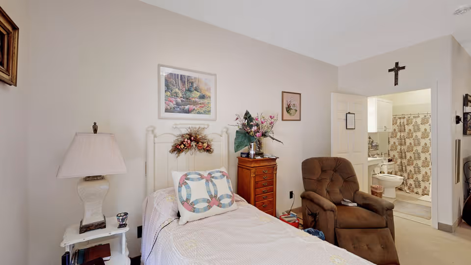 Small bedroom with a twin bed, nightstand and lamp, wooden dresser, recliner chair, and an open doorway leading to a bathroom.