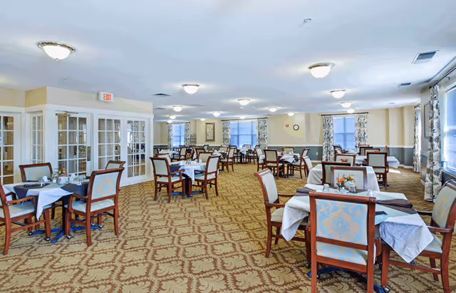 A spacious senior living dining room with multiple tables covered in white tablecloths and set with chairs featuring light blue patterned upholstery. The room has beige walls with white trim, large windows with floral curtains, and a patterned carpet. Ceiling lights illuminate the area, and there are double glass doors on one side.