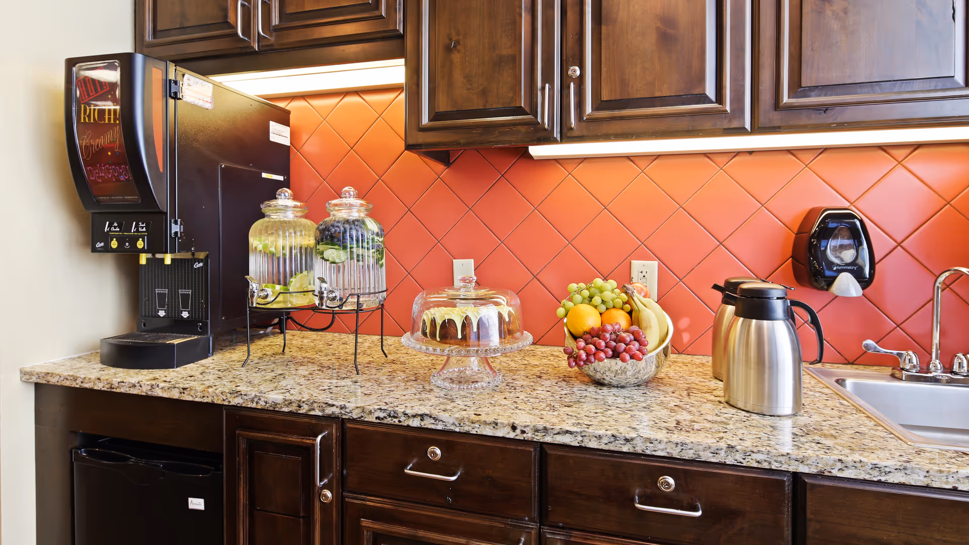 A kitchen countertop with a granite surface featuring a beverage dispenser, two glass containers with infused water, a cake under a glass dome, a bowl of assorted fresh fruit, a stainless steel coffee carafe, and a sink with a faucet. The backsplash is made of red diamond-shaped tiles, and there are dark wooden cabinets above and below the countertop.