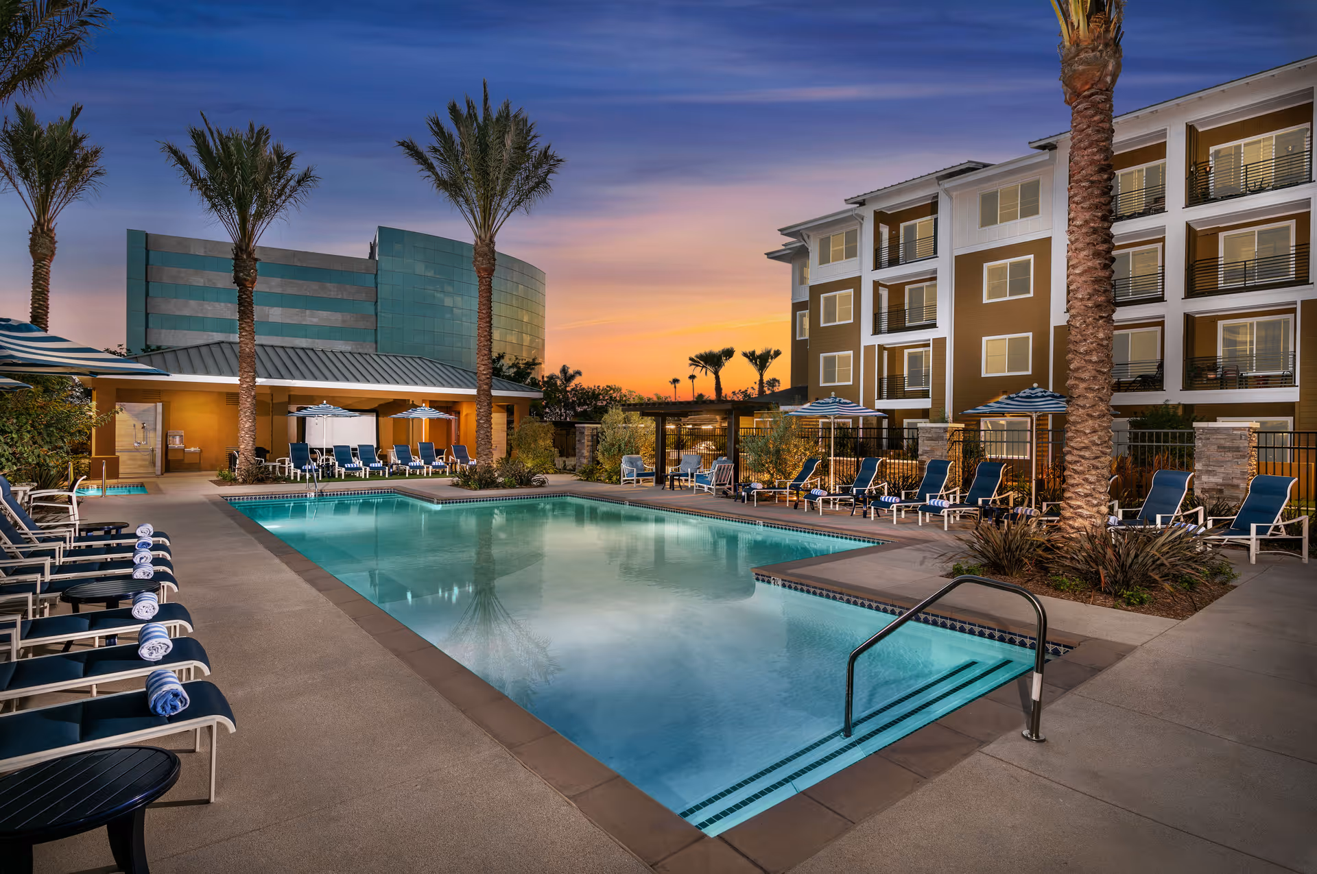 Outdoor swimming pool area at sunset with lounge chairs lined up on both sides, palm trees, umbrellas, and a multi-story building in the background.