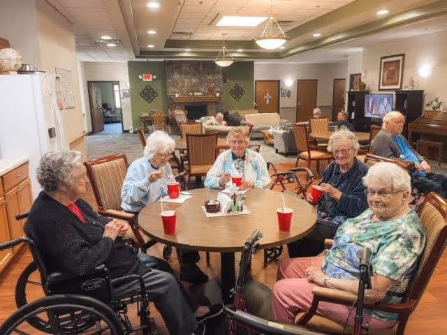 A group of elderly individuals sitting around a round table in a common area of a senior living facility. They are enjoying snacks and drinks from red cups. The room has wooden flooring, comfortable chairs, a fireplace, and a television in the background. The atmosphere appears warm and social.