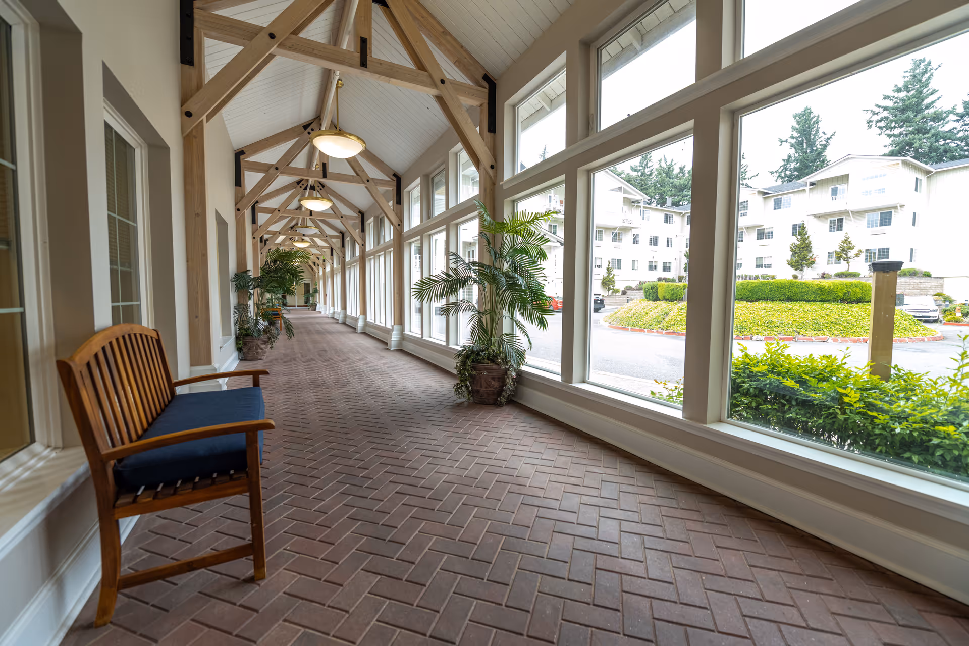 A long indoor corridor with large windows on one side showing an outdoor view of buildings and greenery. The corridor has a brick floor, wooden beams on the ceiling, hanging lights, potted plants, and a wooden bench with a blue cushion.